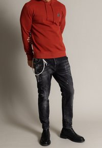 Röd huvtröja med en framficka och en logotypapplikation, som kombineras med slitna svarta jeans och svarta ankelskor. Texturerad, avslappnad outfit.