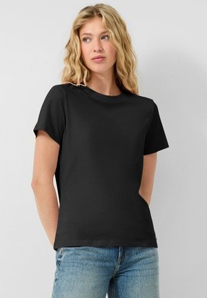 Junge Frau mit welligem blondem Haar, die ein schlichtes schwarzes T-Shirt mit kurzen Ärmeln und blaue Jeans trägt, steht vor einem weißen Hintergrund.