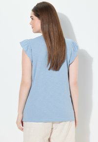 Camiseta de manga corta azul claro con hombros fruncidos, hecha de una tela suave. La parte de atrás es lisa y tiene un corte relajado.