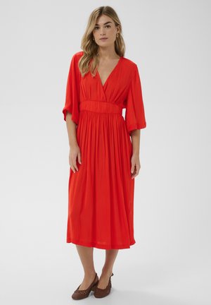 Žena stojaca v jasne červenom midi šatách s hlbokým výstrihom do V, širokými rukávmi a hnedými nízkymi pletenými topánkami na jednoduchom pozadí.