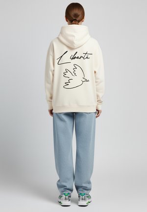 Felpa color crema con una grafica di un uccello e la parola "Liberté" sulla schiena; abbinata a pantaloni leggeri blu chiaro con texture.