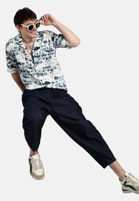 Camicia a maniche corte con bottoni, caratterizzata da una stampa astratta blu e bianca, abbinata a pantaloni in denim scuro e sneakers in stile retrò.