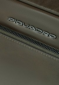 Accessorio in pelle Piquadro verde oliva scuro, caratterizzato da un logo argentato e una cerniera con denti neri lungo il bordo. Texture liscia.