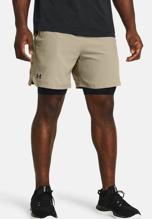 Beige athletic shorts with a smooth texture, featuring side pockets and a black logo. Worn over black compression shorts with athletic shoes.