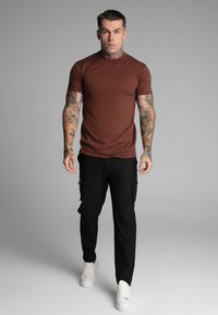 Brown short-sleeve fitted shirt, black cargo pants with side pockets, and white sneakers. Smooth fabric with no visible patterns or logos.
