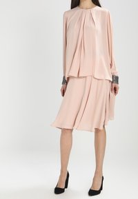 Lyserød kjole med flydende design, plisseret bodice, lange ærmer og udsmykkede manchetter. Kombineret med sorte højhælede sko.