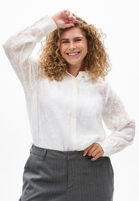 Bílá průsvitná blůza s květinovým vzorem, s knoflíkovým zapínáním vpředu a dlouhými rukávy, spárovaná se šedými pruhovanými kalhotami.