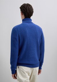 Maglione azzurro lavorato a maglia con colletto alto, polsini e orlo a coste; visto da dietro, abbinato a pantaloni di colore chiaro.