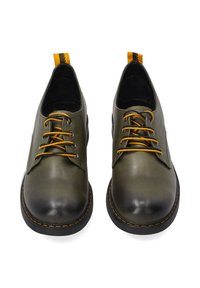Groene leren schoenen met een zwarte neuskap, gele veters en een zwarte en gele trekband achteraan. Stikdetails zijn zichtbaar.