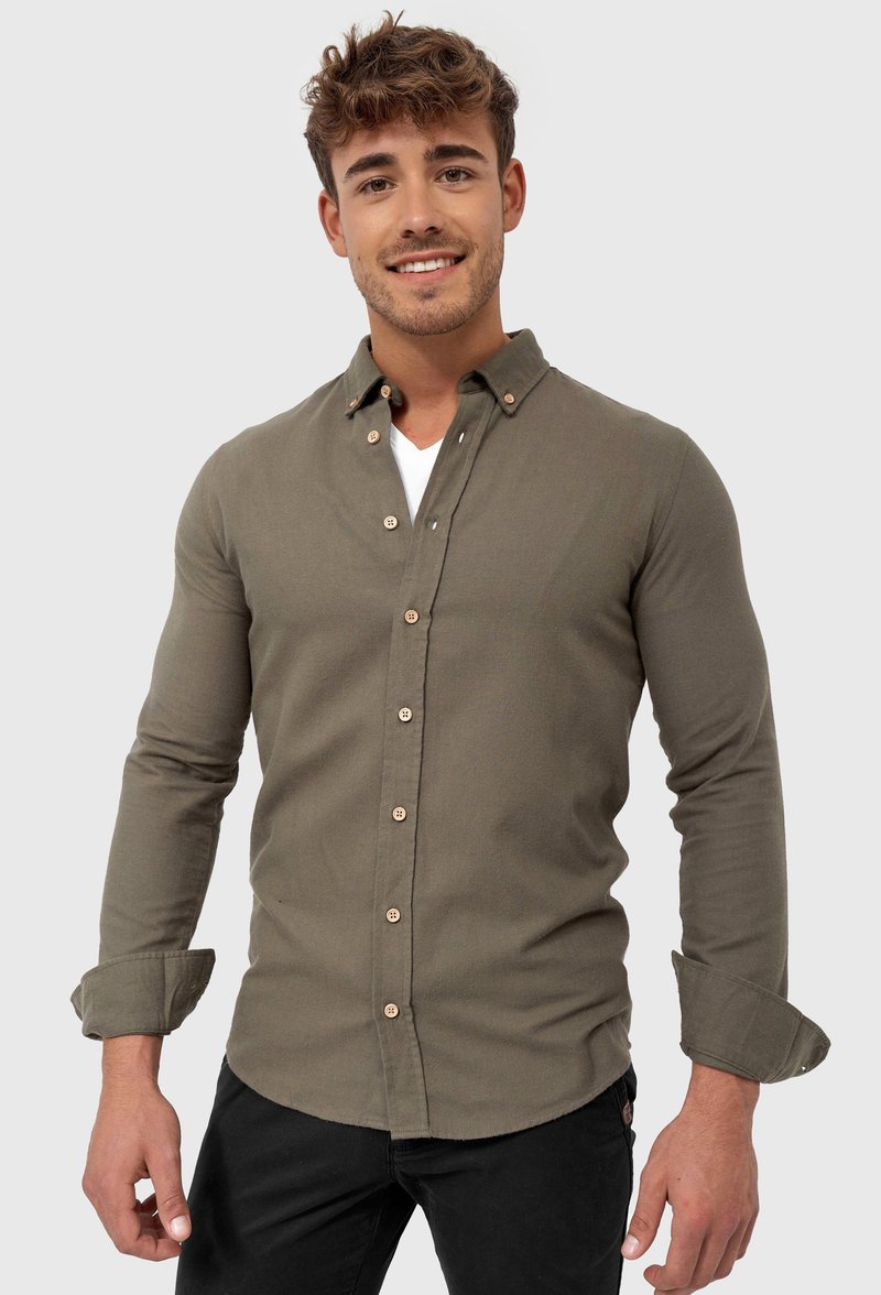 Camisa de botones verde oliva con una textura suave, que presenta un cuello, detalles de botones y mangas enrolladas. Usada sobre una camiseta blanca.