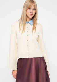 Lys beige cardigan med ribbet tekstur, rund hals, og frontknapper. Kombinert med en blå skjorte med krage og et burgunder skjørt.