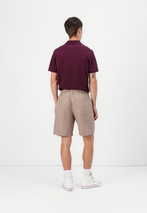 Homme portant un polo bordeaux, un short beige avec taille élastique, des chaussettes blanches et des baskets montantes blanches, debout face à un mur blanc.