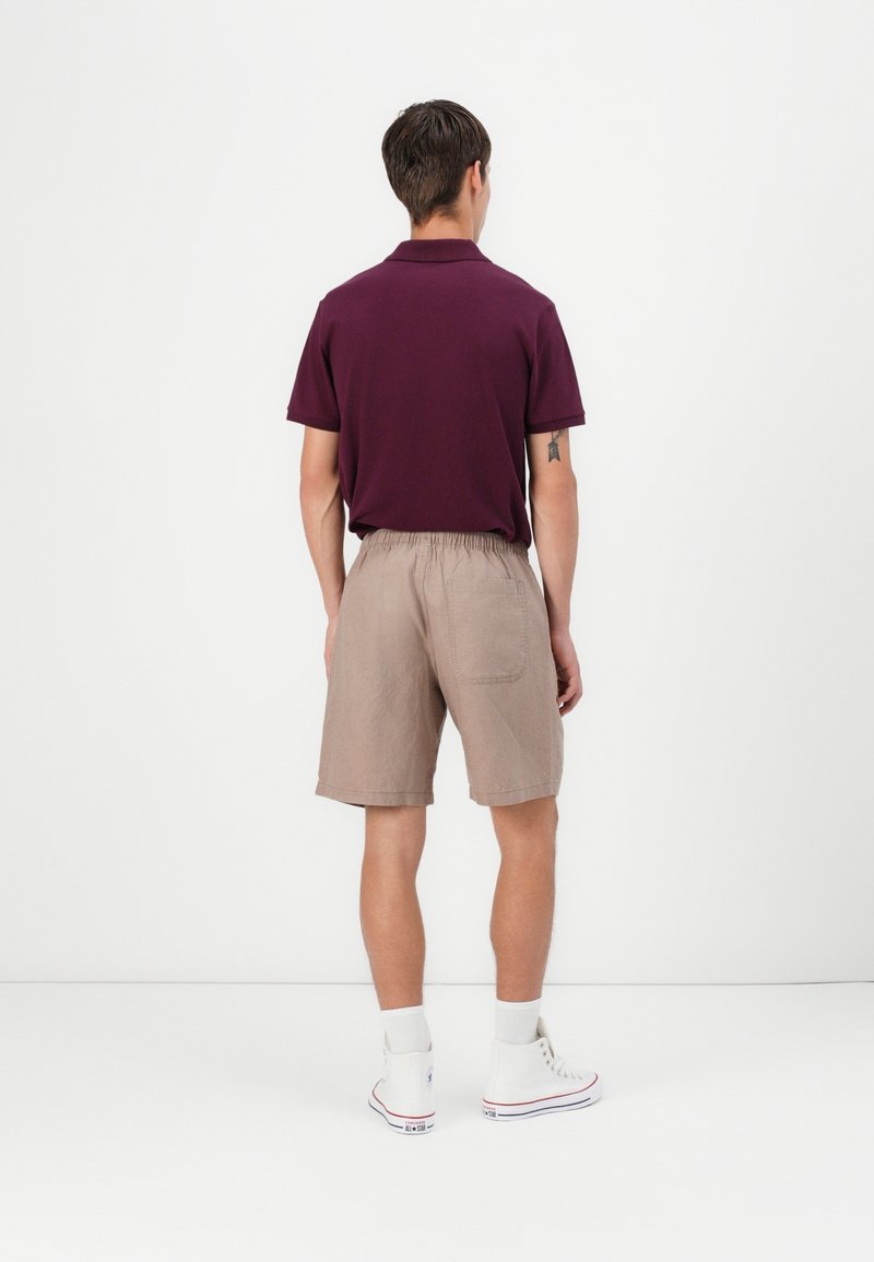 Homme portant un polo bordeaux, un short beige avec taille élastique, des chaussettes blanches et des baskets montantes blanches, debout face à un mur blanc.