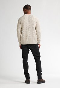 Kreativ kabelstickad tröja med ribbad mudd och fåll, i kombination med svarta slim-fit jeans och bruna ankelskor. Visad bakåtvänd.