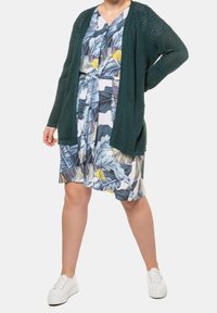 Žena oblečená v modrožlutých šatech s tropickým vzorem, tmavě zeleném kardiganu s otevřeným pletením a bílých teniskách, stojící s jednou rukou na kyčli.