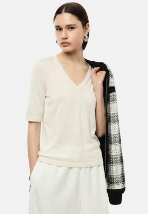 Giovane donna con capelli corti e scuri indossa una maglia color crema con scollo a V e pantaloni coordinati, tenendo una giacca a quadri bianco e neri sulla spalla.