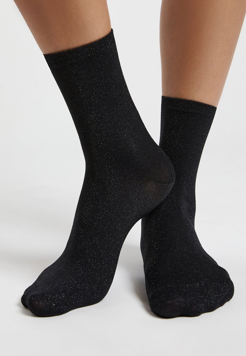 Schwarze Knöchelsocken aus dehnbarem Stoff mit glitzernder Textur. Sie verfügen über einen gerippten Bund und eine glatte Oberfläche.