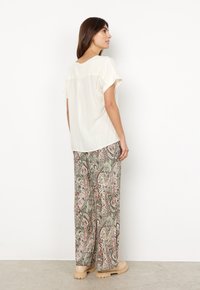 Kvinde set fra siden iført en kortærmet cremegul bluse, bredslåede bukser med paisley-mønster og beige klodsede støvler mod en ensfarvet væg.