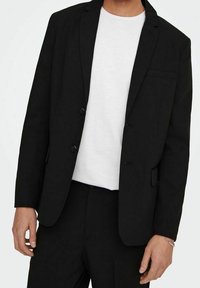 Czarna marynarka charakteryzuje się dopasowanym krojem, jednorzędowym fasonem, dwoma guzikami i kieszonką na piersi, zestawiona z gładką białą koszulką pod spodem.