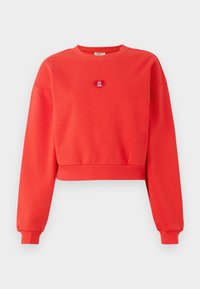 Sweatshirt rouge court en tissu doux, avec des manches longues et un ourlet côtelé. Un petit patch en forme de cœur sur le devant affiche le texte "LE CLUB VIP."