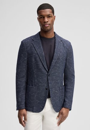 Mann trägt ein dunkelblau strukturiertes Sakko über einem schwarzen Shirt und weißen Hosen, steht vor einem einfarbigen hellgrauen Hintergrund.