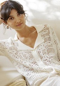 Femme aux cheveux foncés portant des boucles d'oreilles anneaux argentées et une chemise blanche à col en maille ouverte avec boutons, reposant sa tête sur sa main.