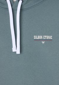 Sudadera verde de algodón con cordones blancos, un pequeño logotipo "SILBON ETHNIC" en blanco y rojo, y textura suave.