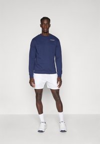 Marineblauwe sweatshirt met lange mouwen, ronde hals, ribgebreide manchetten en logo op de borst, gecombineerd met witte sportshorts en sneakers.