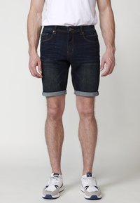 Pantalones cortos de mezclilla oscura con dobladillo vuelto, que presentan un diseño clásico de cinco bolsillos y costuras en naranja contrastante. Llevados con una camiseta blanca y zapatillas.