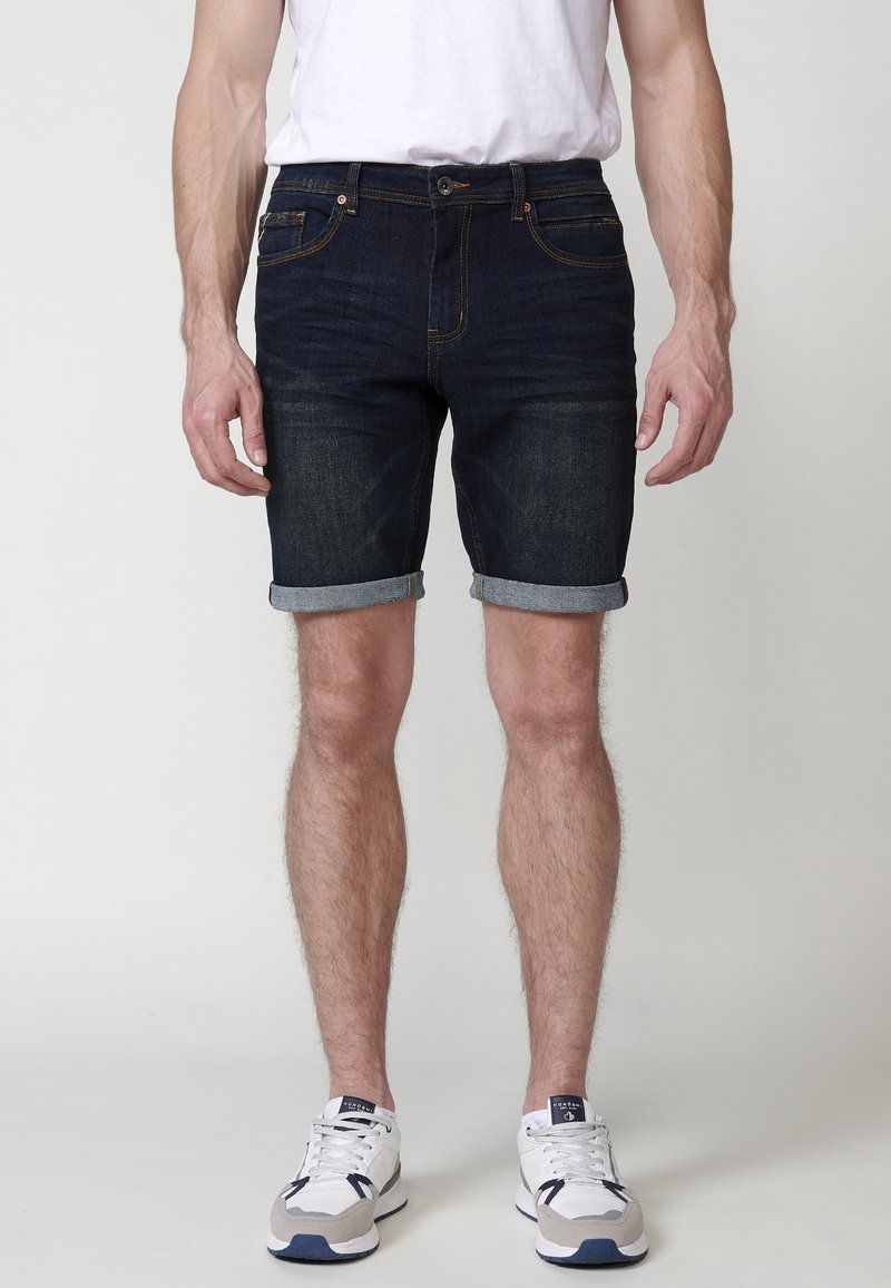 Pantalones cortos de mezclilla oscura con dobladillo vuelto, que presentan un diseño clásico de cinco bolsillos y costuras en naranja contrastante. Llevados con una camiseta blanca y zapatillas.
