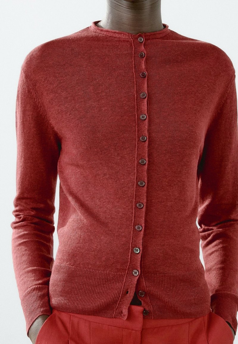 Personne portant un cardigan rouge ajusté boutonné avec poignets et ourlet côtelés, associé à un pantalon rouge, mains dans les poches, sur un fond uni.