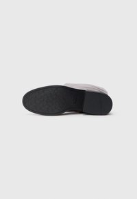 Suela de goma negra de una bota con un patrón de agarre texturizado. El material superior parece ser suave y posiblemente de cuero, en un color claro.