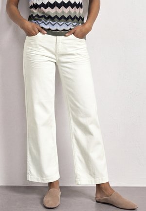 Person iført hvide bukser med vidde ben, flerfarvet zigzag-mønster top, hænder i lommerne og beige slip-on flade sko, der står mod en væg.