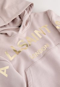 Lichtroze hoodie met gouden metallic "ALLSAINTS LONDON"-tekst, met een voorzak en capuchon, plat neergelegd op een witte ondergrond.