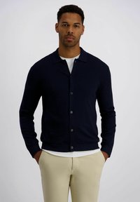 Marineblauer Cardigan mit Schalkragen, Frontknöpfen und langen Ärmeln. Kombiniert mit einem weißen Hemd und beigen Hosen mit glatter Textur.