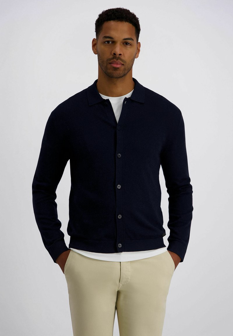 Marineblauer Cardigan mit Schalkragen, Frontknöpfen und langen Ärmeln. Kombiniert mit einem weißen Hemd und beigen Hosen mit glatter Textur.