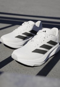Weiße Sportsneaker mit einer strukturierten Oberfläche, die an den Seiten drei auffällige schwarze Streifen und eine schlanke, gebogene Sohle aufweisen.