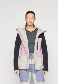 Schneejacke in einem Mix aus Lavendel, Beige und Schwarz. Verfügt über eine Kapuze, Fronttaschen und strukturierte Stoffe. Kombiniert mit helllila Hosen.