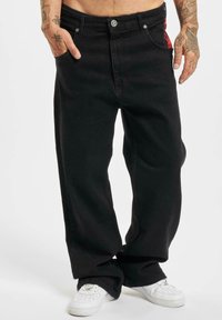 Schwarze weite Jeans mit glatter Textur, ausgestattet mit vorderen Taschen, einem Knopfverschluss und einem roten Akzent auf der linken Seite. Darunter weiße Sneakers.