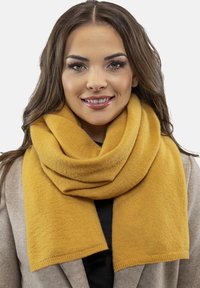 Musztardowo-żółty wełniany szalik, miękka faktura, elegancko owinięty wokół szyi, nałożony na lekki płaszcz, o gładkich krawędziach i bez widocznych wzorów.
