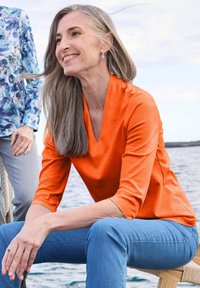 Oranje V-hals top met driekwart mouwen, gemaakt van een gladde stof, gecombineerd met blauwe spijkerbroek. Heeft natuurlijke, vloeiende haren en turquoise oorbellen.