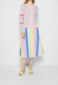 Pastellfärgad cardigan med rutmönster och randiga ärmar, tillsammans med en färgglad randig klänning. Skorna är gröna, fyrkantiga slides.