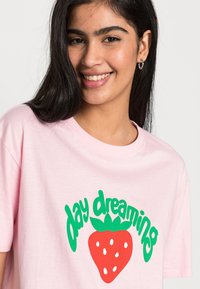T-shirt de algodão rosa com o texto "sonhando acordado" em verde e uma ilustração de morango vermelho com sementes brancas, gola redonda e mangas curtas.