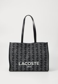 Geantă mare rectangulară din material negru, cu un model geometric. Include două mâneri rezistente negre și un logo alb "LACOSTE 1933".