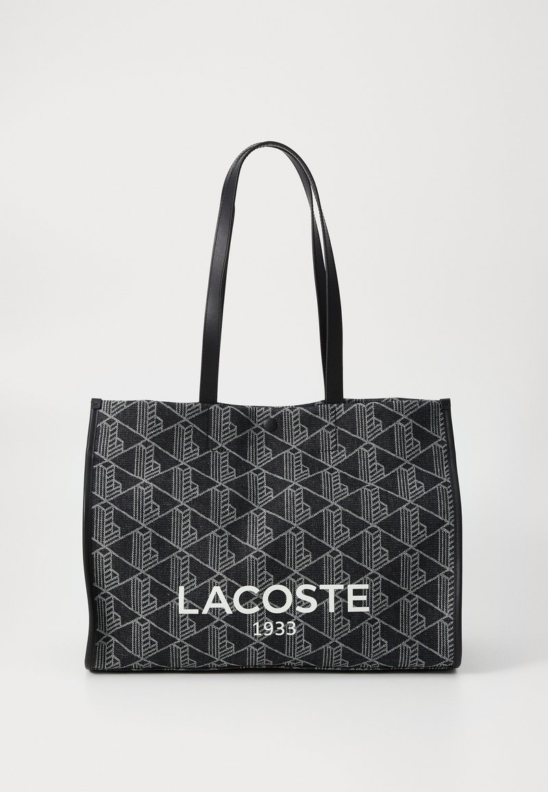 Geantă mare rectangulară din material negru, cu un model geometric. Include două mâneri rezistente negre și un logo alb "LACOSTE 1933".