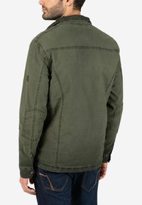 Olivgrüne, leichte Jacke mit Kragen, Seitentaschen und geknöpften Manschetten, die eine glatte Textur und genähte Details an den Nähten aufweist.