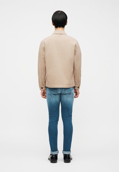 Άτομο στέκεται με την πλάτη γυρισμένη φορώντας μπεζ μπουφάν, μπλε στενά τζιν με κουμπωμένα μπατζάκια και μαύρα επίσημα παπούτσια μπροστά σε απλό λευκό φόντο.