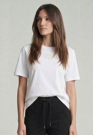 Frau mit langen braunen Haaren, die ein weißes T-Shirt mit einem goldenen Lippen-Design und schwarze Hosen mit Kordelzug trägt, schaut seitlich vor einem schlichten Hintergrund.