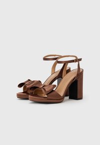 Talons plateformes en satin marron avec un accent en nœud, orteils ouverts, bride de cheville et talon bloc. Matériau texturé avec une finition lisse et brillante.