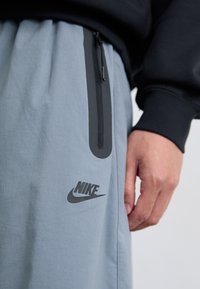 Et nærbilde av en persons hånd som holder livet på lysegrå Nike-bukser med en glidelåslomme og logo synlig.